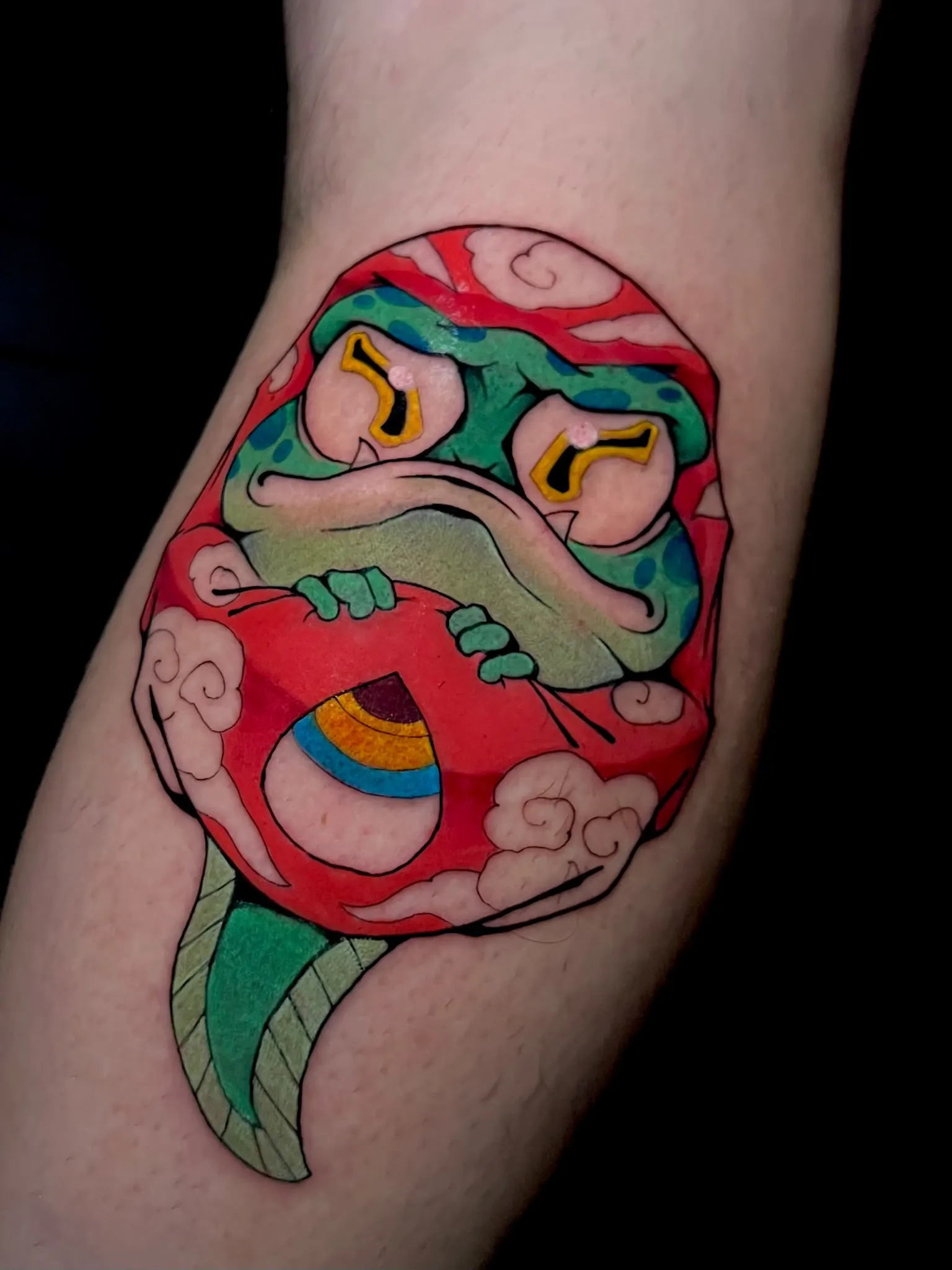Tatuaje Illustrative Color de un Daruma rojo con ojos desiguales sobre nubes y hojas verdes, tatuado en el muslo