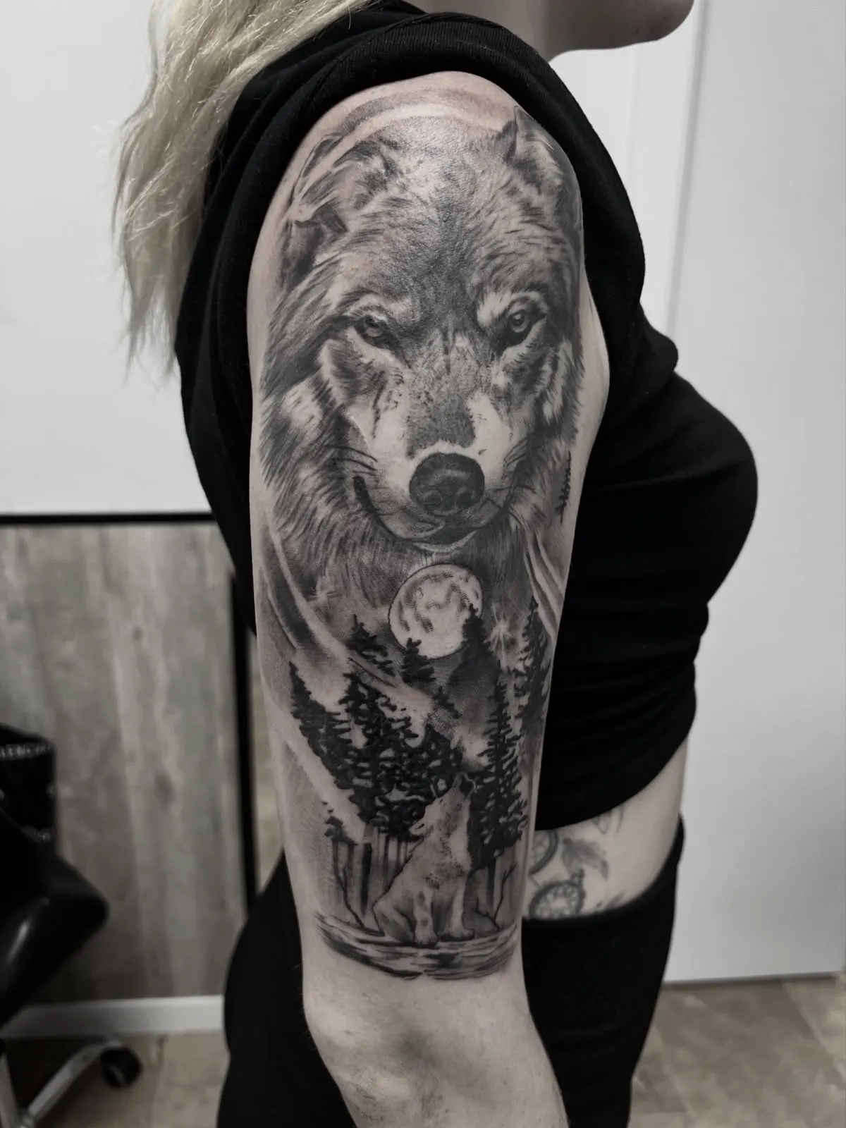 Tatuaje realista en blanco y negro de un lobo aullando con luna y bosque de pinos en el hombro y antebrazo